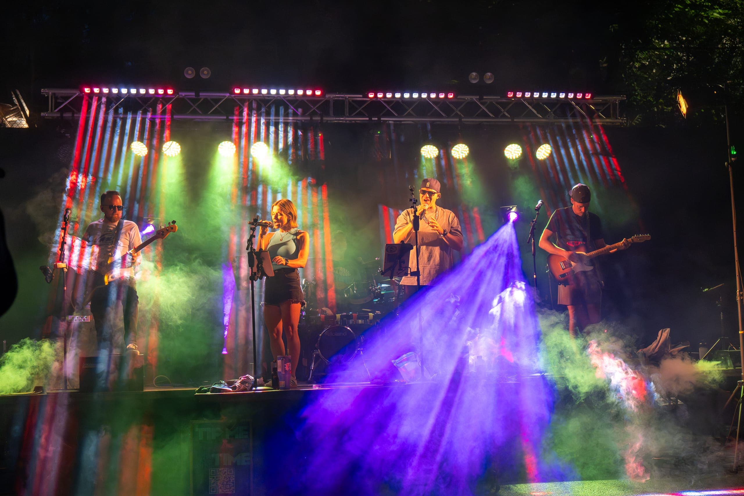 Full band performing on an outdoor stage bathed in colorful lights and fog